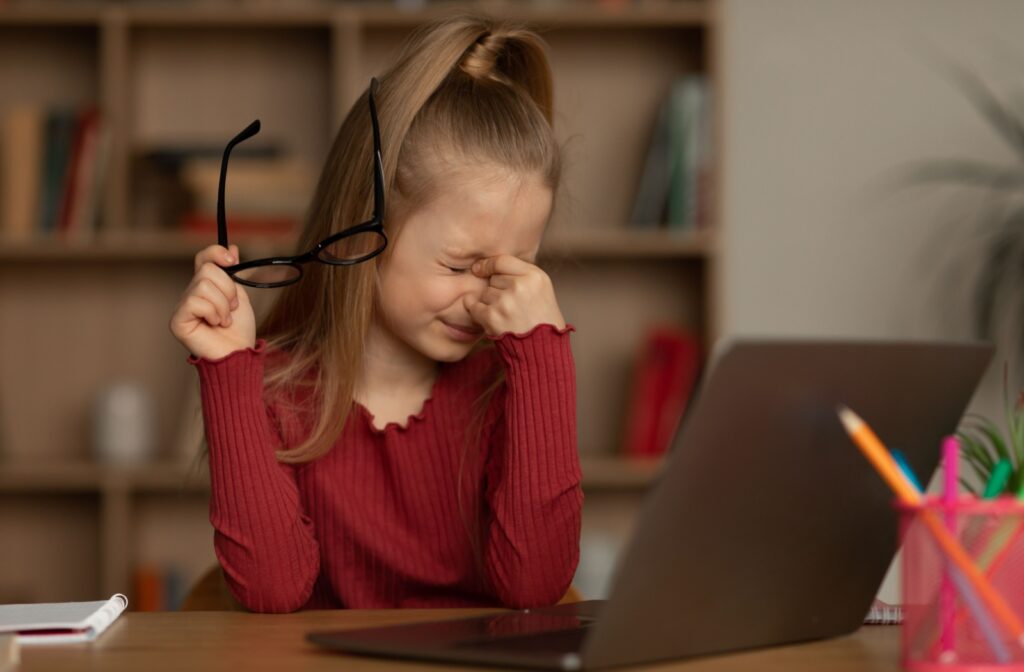 A young child in a red shirt pulls off their glasses to rub their eyes while sitting in front of a laptop