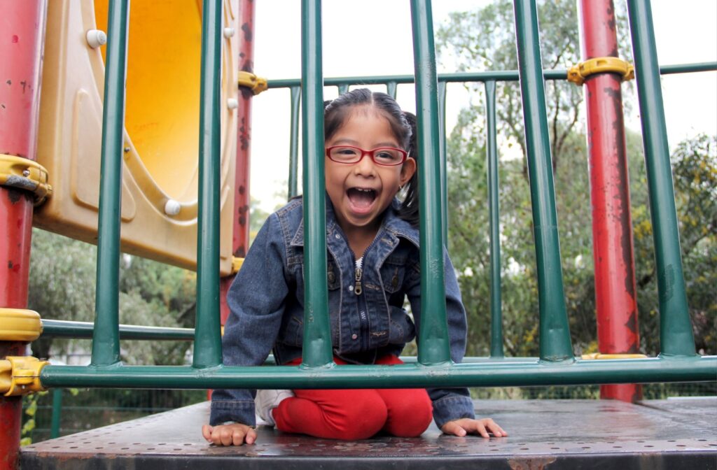 A young child in red glasses laughs while peeking through the bars of playground equipment next to a yellow slide