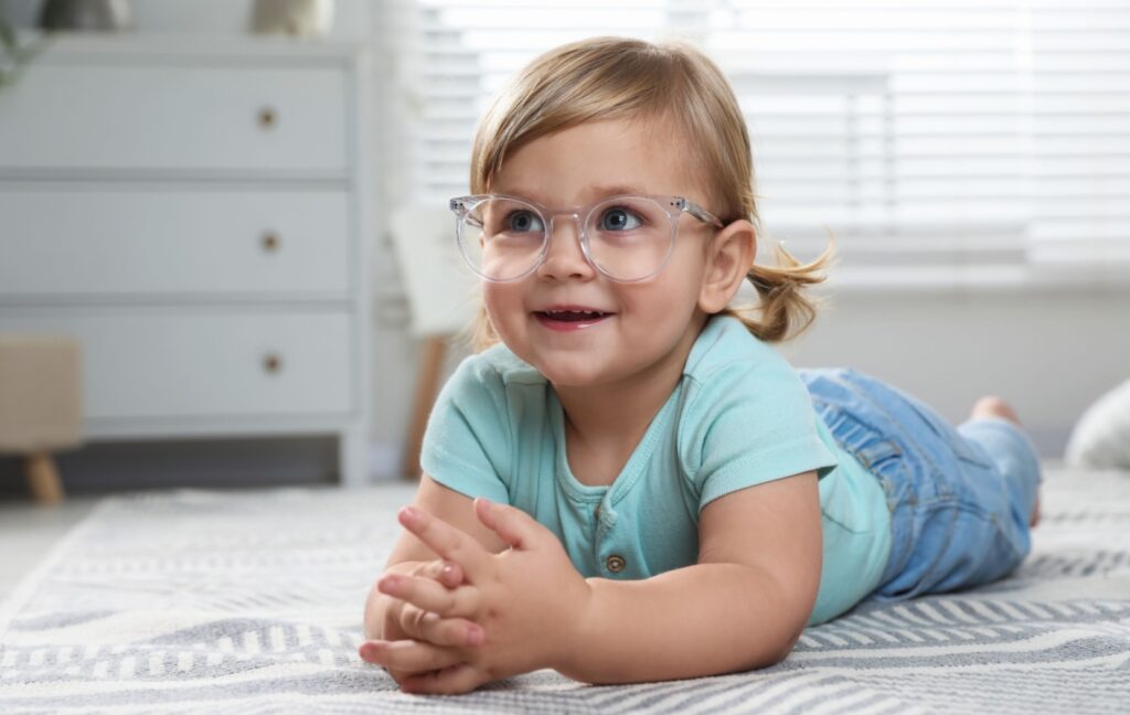 A preschool-aged child lies on the floor and looks into the distance. They are wearing a pair of glasses for myopia correction.